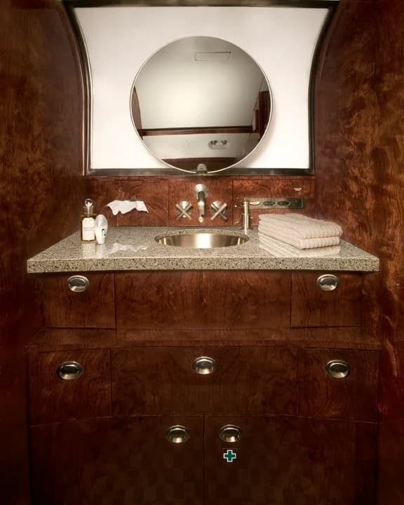 The private jet lavatory features a polished metal sink, a round mirror, and dark wood cabinetry with a granite-like countertop.