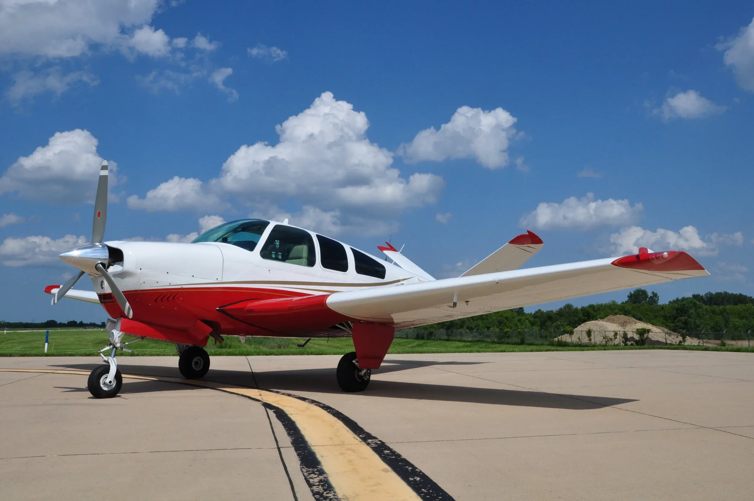 A white and red single-engine propeller aircraft is parked on an asphalt runway under a blue sky with white clouds.