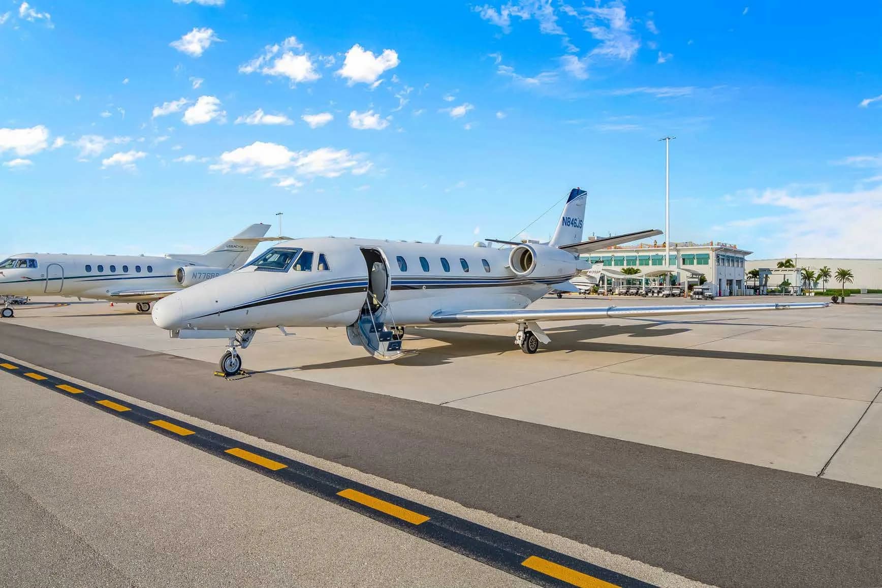 A white Cessna Citation business jet, N846JS, with its main door open, parked on an airport tarmac with other jets and buildings in the background.