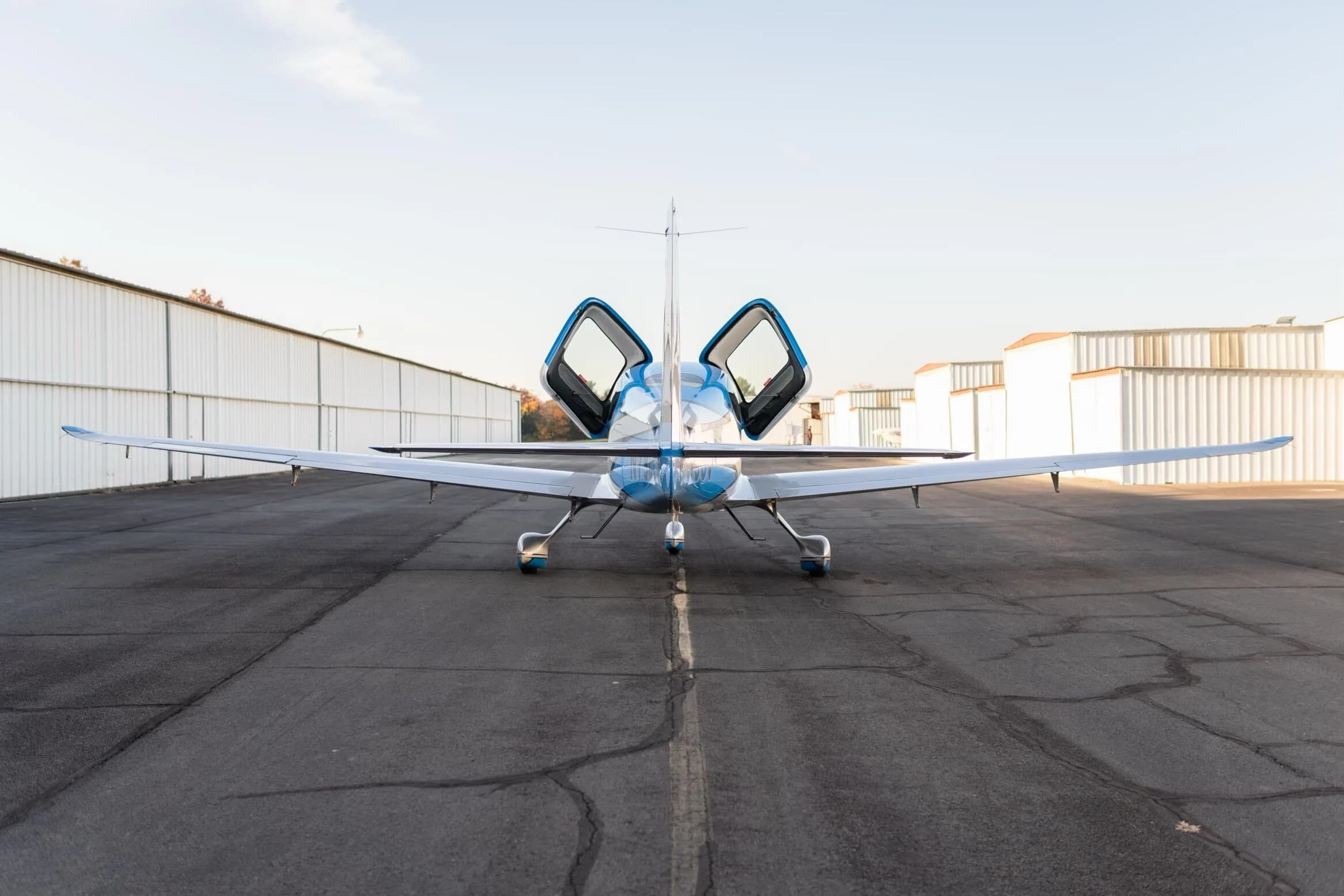 A rear view of the blue and silver Cirrus SR22T GTS aircraft, registration N79CV, with its gull-wing doors open, parked on an airfield.
