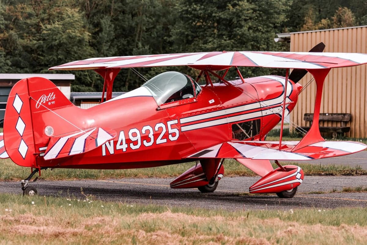 A red and white Pitts S-1T biplane with registration N49325 parked on the grass in front of a hangar.