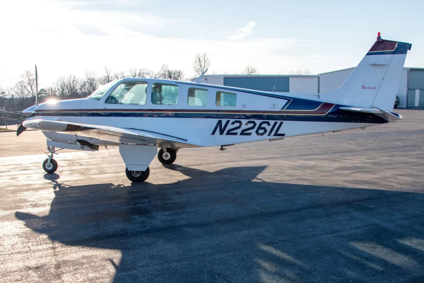 A white Beechcraft Bonanza N2261L with blue and dark blue stripes parked on an airfield with sun glare.