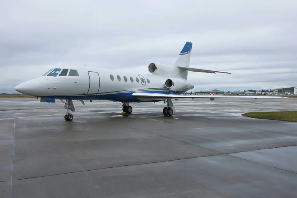 A white business jet with blue stripes parked on a wet tarmac under a cloudy sky.