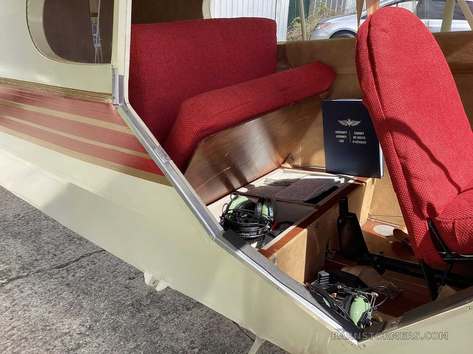 The aircraft's interior shows red fabric seats, a wooden storage area, and a flight logbook with a headset.