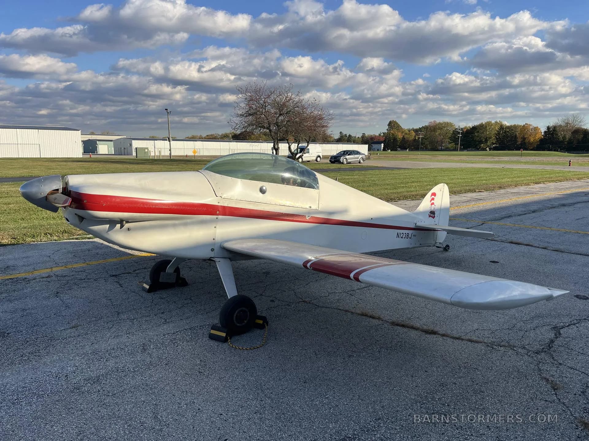 A white and red single-engine aircraft with a propeller, parked on an asphalt apron under a partly cloudy sky during the day.