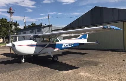 A white single-engine propeller aircraft with blue and red stripes, registration N61557, parked on an airfield in front of a hangar.