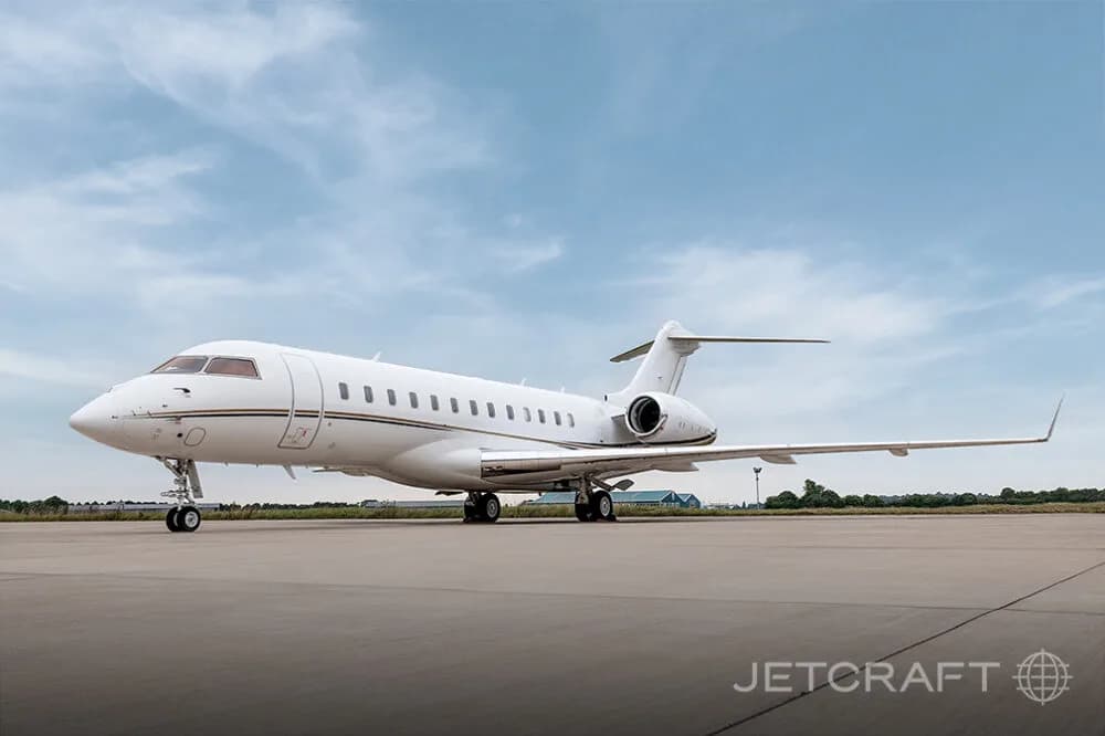 A white Bombardier Global business jet with gold and black stripes parked on an airfield under a partly cloudy sky.