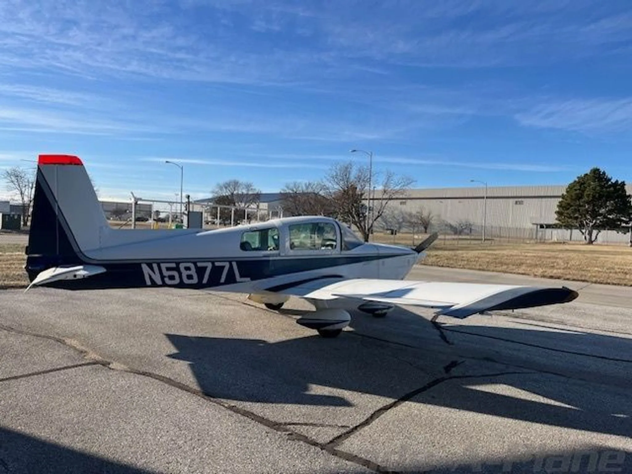 A white and blue Grumman AA-5B Tiger aircraft (N5877L) parked on a tarmac with buildings and trees in the background.