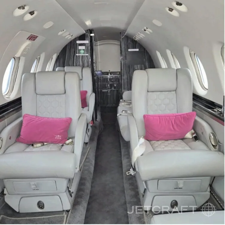 A view down the aisle of a private jet cabin, showing light grey seats with pink pillows.