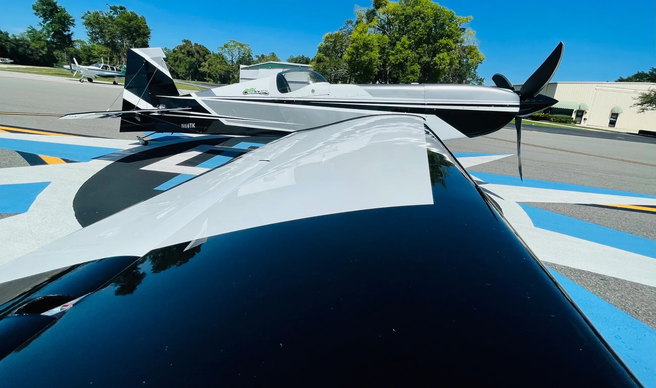 A black and white aerobatic aircraft (N66TK) parked on a patterned tarmac, viewed over its wing.