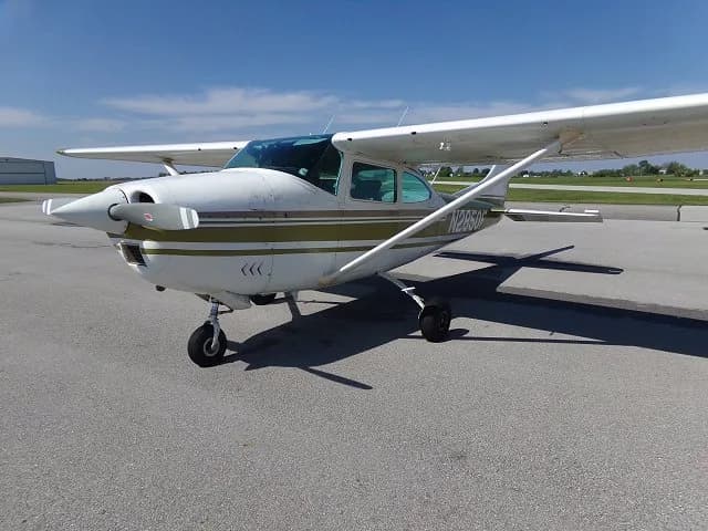 A front three-quarter view of a white Cessna 182 with olive green and brown stripes, registration N2850F, parked on an asphalt tarmac.