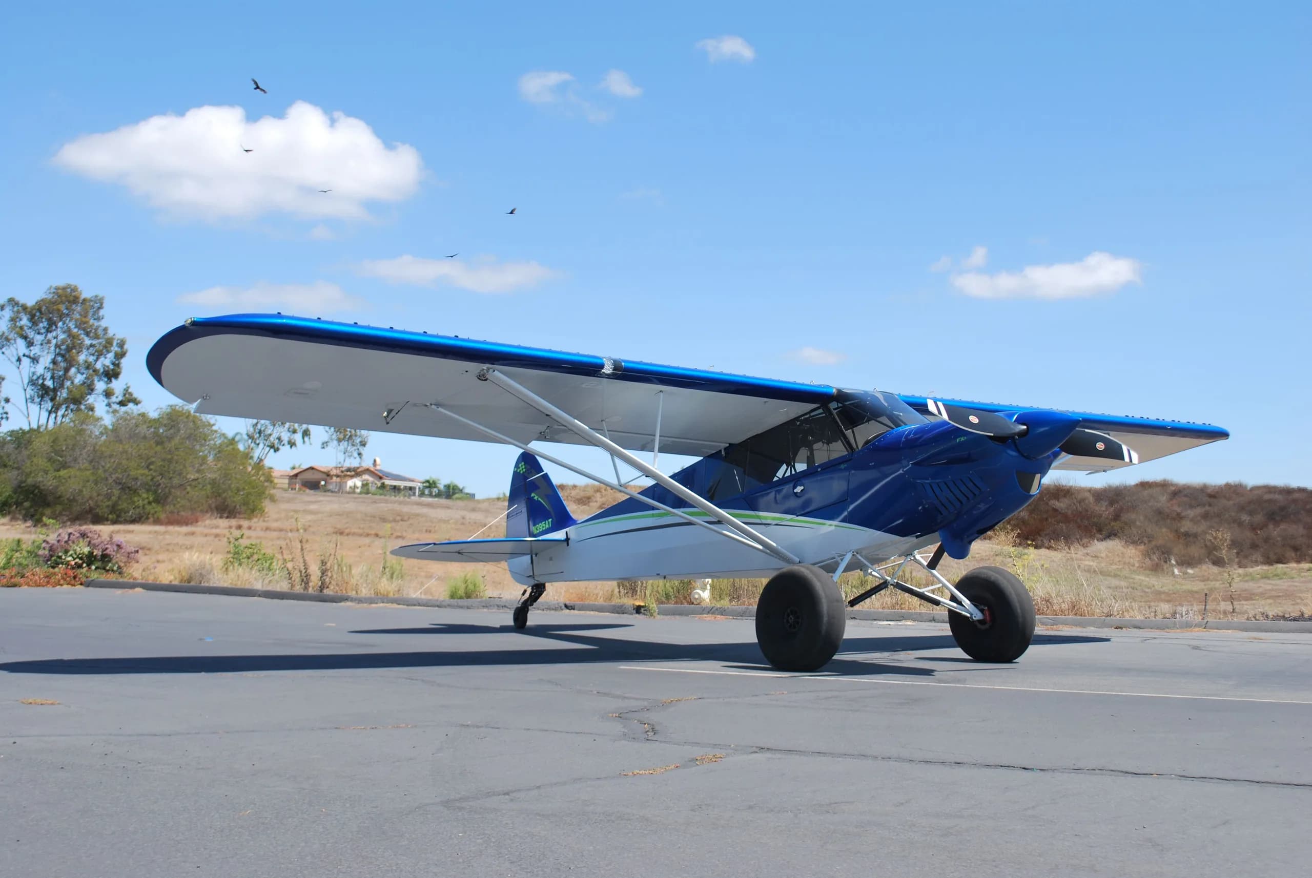 A blue and white Carbon Cub FX3 aircraft with large tires parked on an asphalt ramp with a hilly, dry landscape in the background.