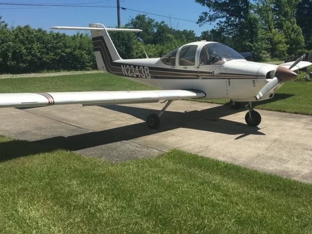 A Mooney M20C aircraft with a white and brown paint scheme is parked on a concrete surface.