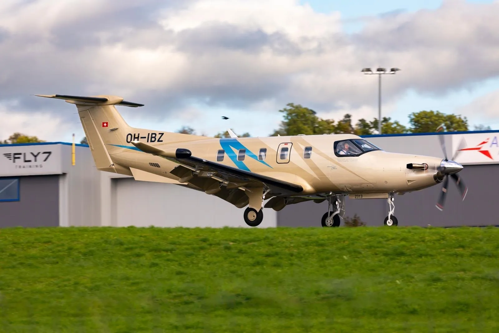A beige Pilatus PC-12 NGX aircraft with blue accents and registration OH-IBZ is seen on a grassy field, possibly taking off.
