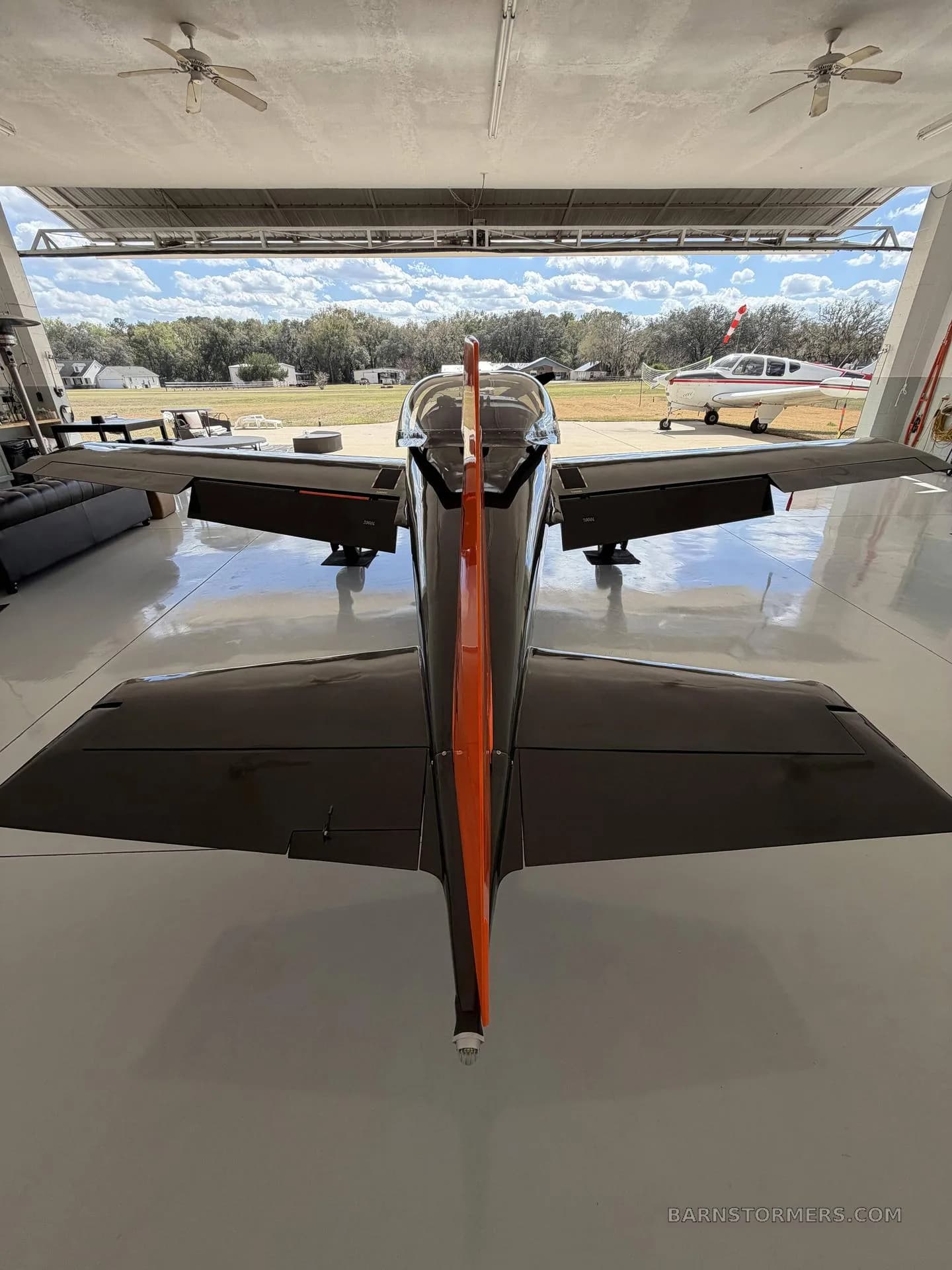 A rear view of the black and orange single-engine aircraft with a prominent orange vertical stabilizer, looking out of a hangar.