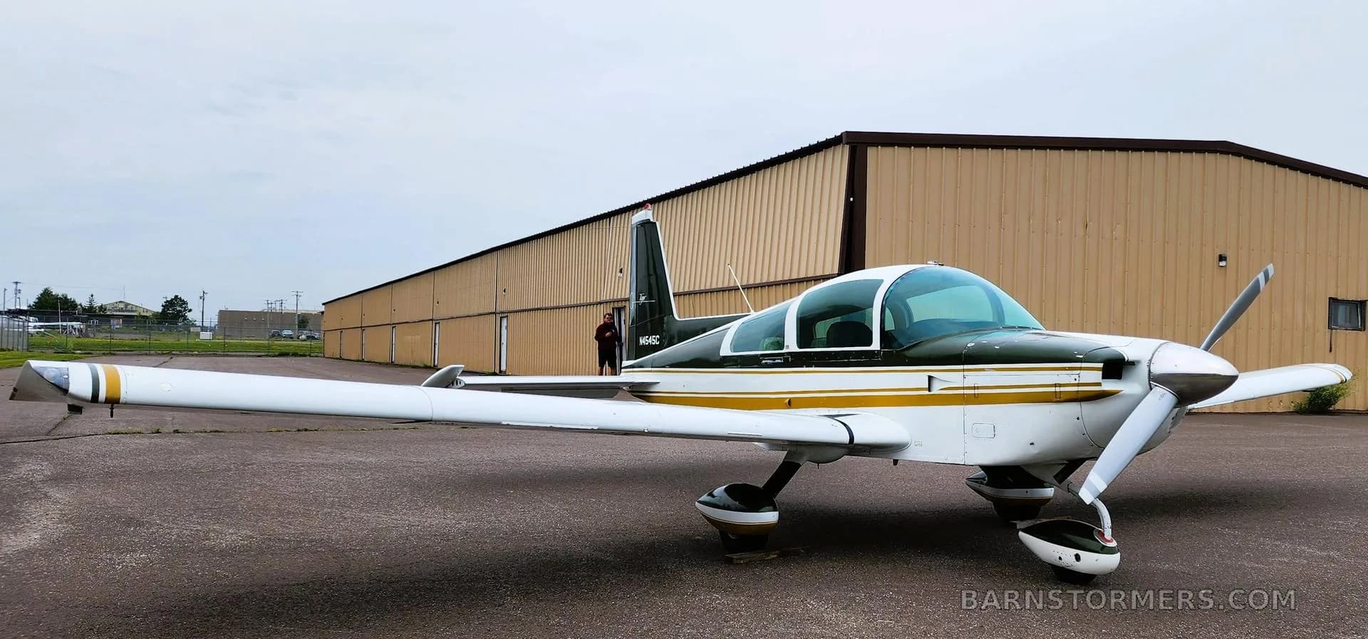 A white and dark green Grumman Tiger aircraft with gold stripes, parked on an asphalt ramp next to a hangar.