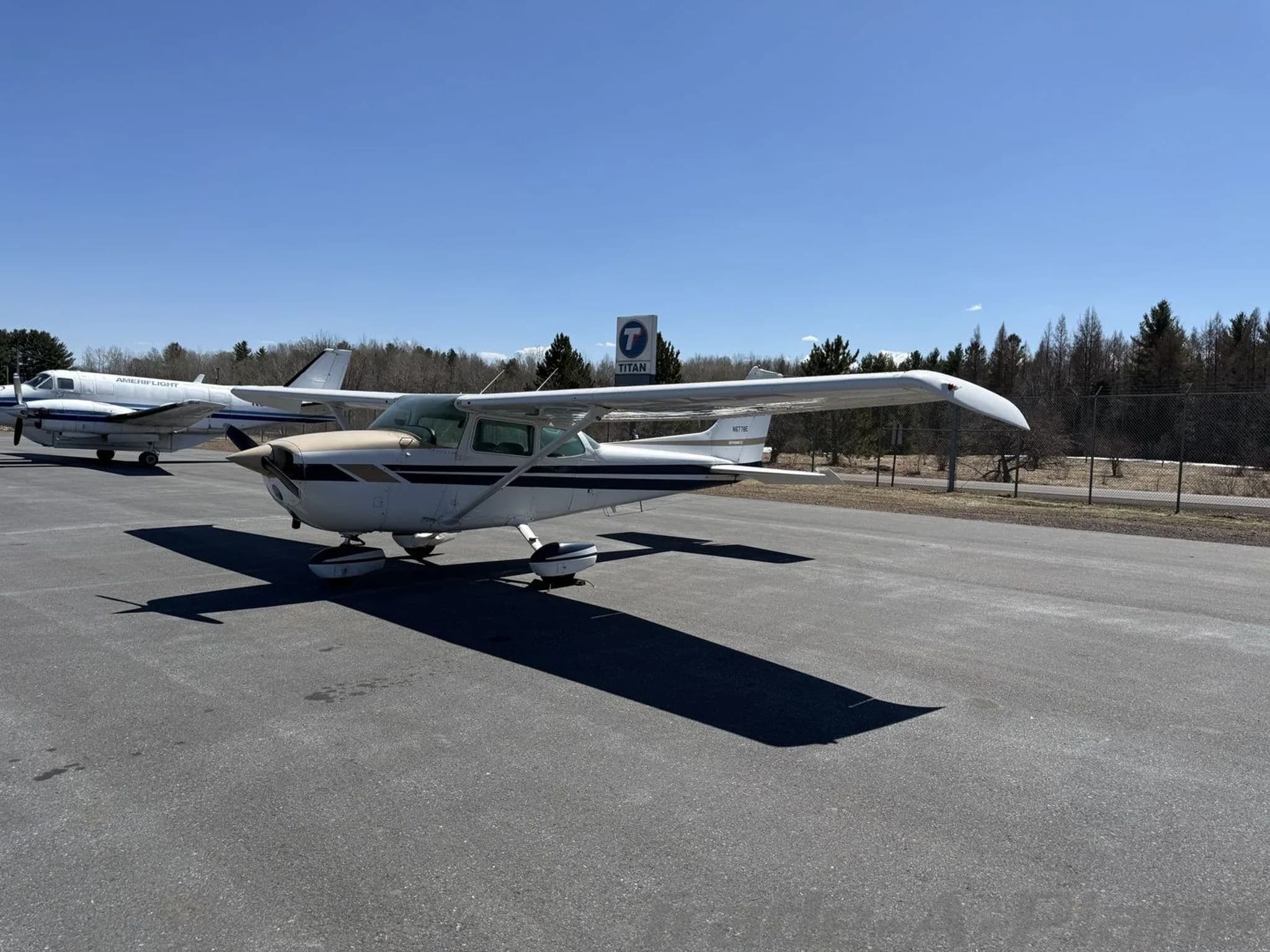 A white and tan Cessna 172 aircraft with blue stripes parked on an asphalt apron under a clear blue sky.