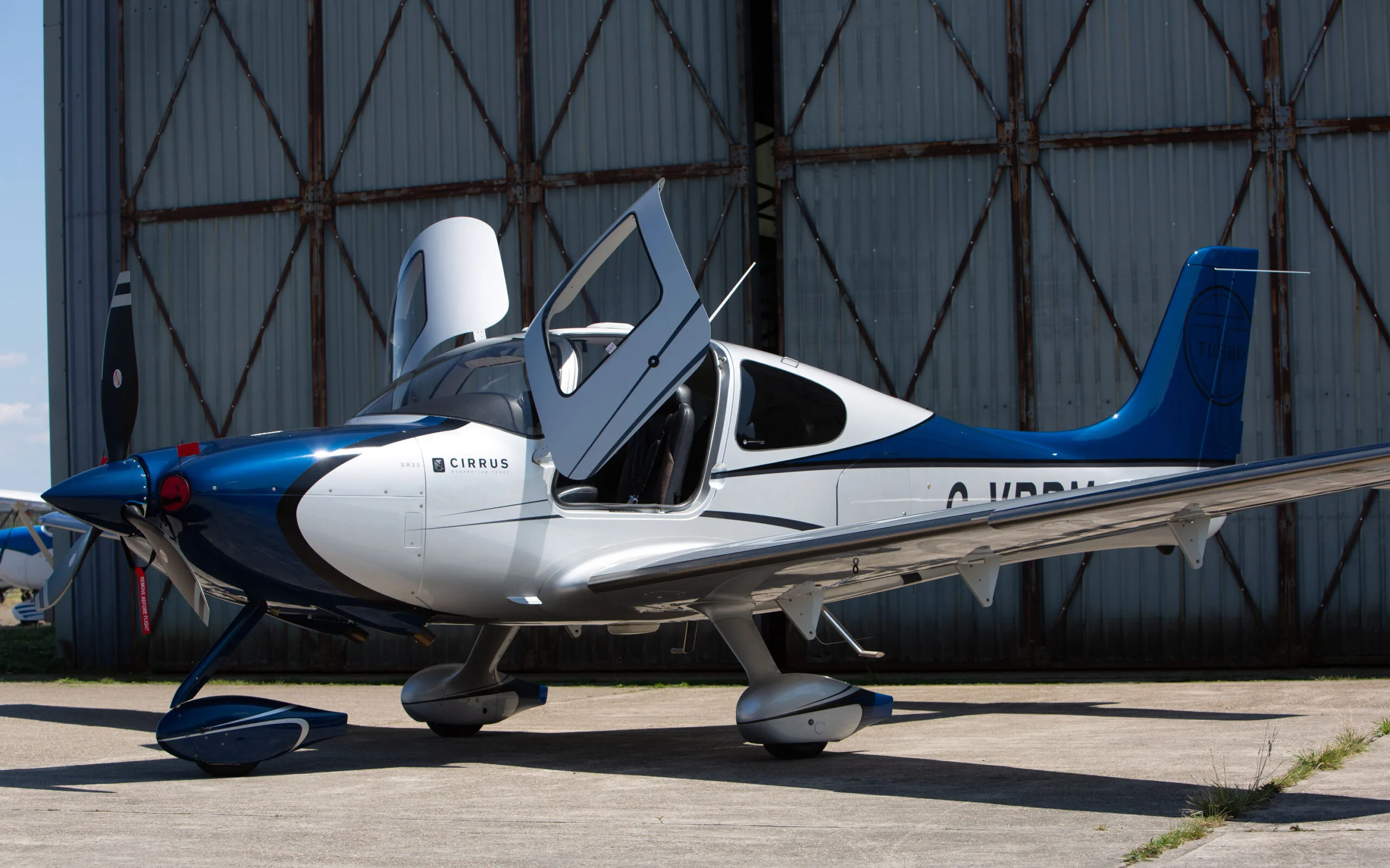 A blue and white Cirrus SR22 aircraft with registration G-VBPM and its gull-wing doors open, parked on an airfield apron.