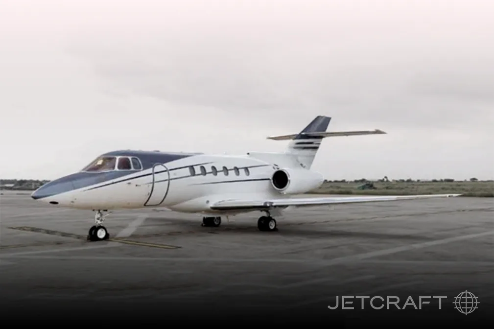 A white and blue business jet, likely a Hawker, is parked on an asphalt tarmac under an overcast sky.
