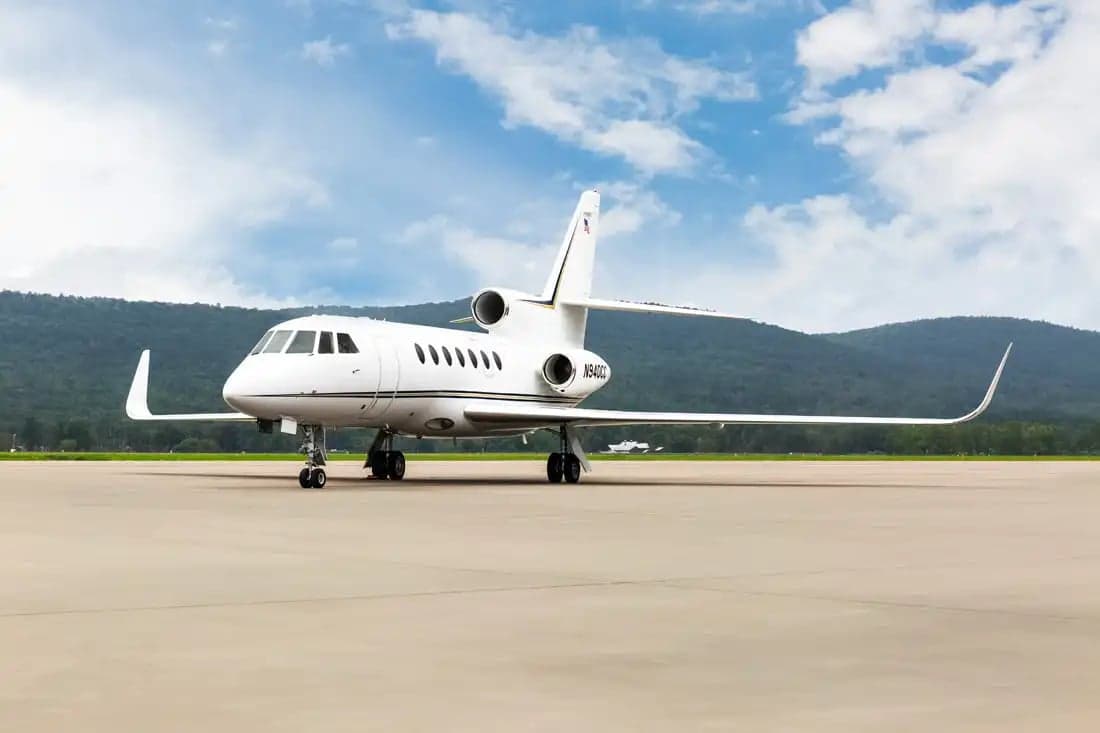 A Falcon jet sits on the tarmac with mountains in the background.