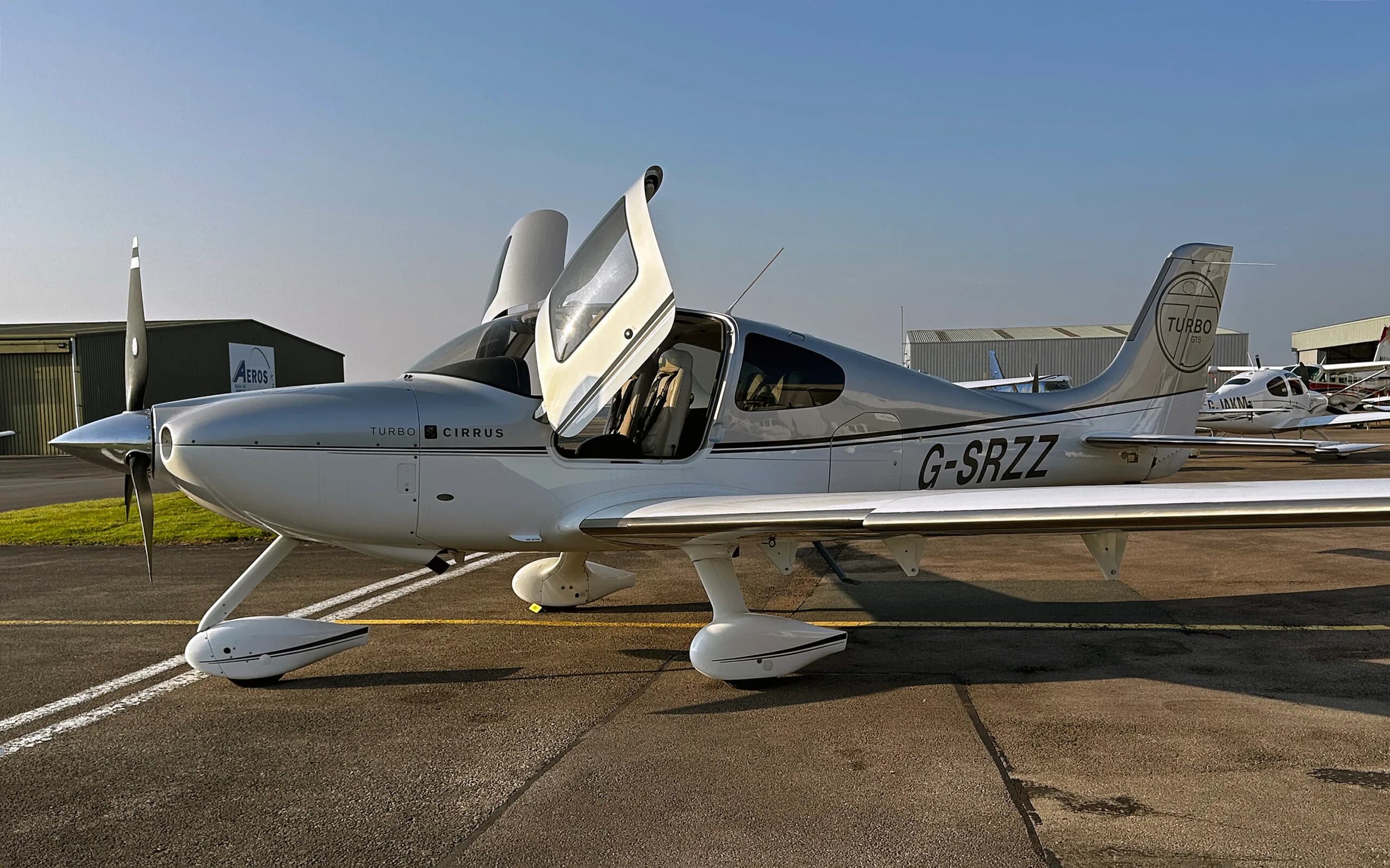 A silver and white Cirrus SR22 Turbo aircraft with registration G-SRZZ and open doors, parked on an airfield.