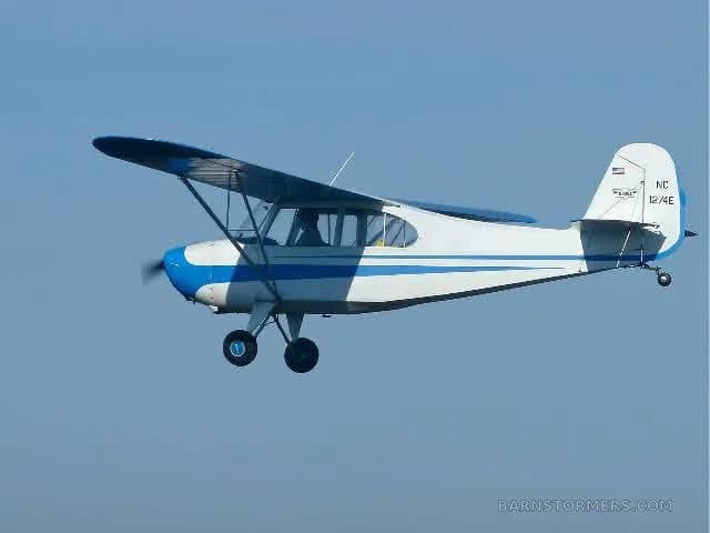 A white and blue single-engine high-wing aircraft, registration NC 1274E, flying in a clear blue sky.