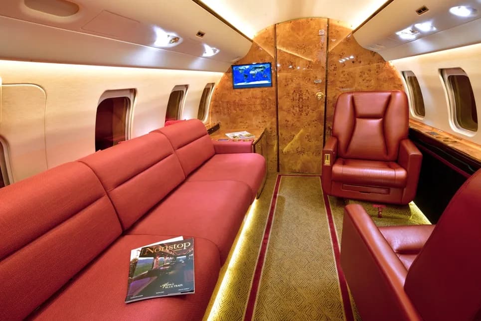 The aircraft cabin interior featuring a long red sofa, a single red armchair, and burled wood walls.