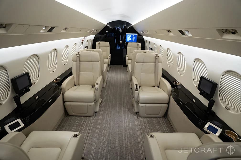 The luxurious interior cabin of a business jet features light beige leather seats, dark wood accents, and individual entertainment screens.