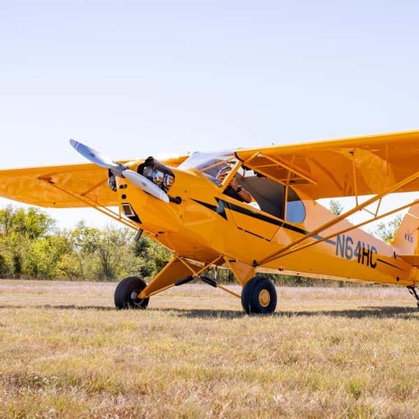 A yellow Legend Cub aircraft, registration N64HC, is parked on a grassy field under a clear blue sky.