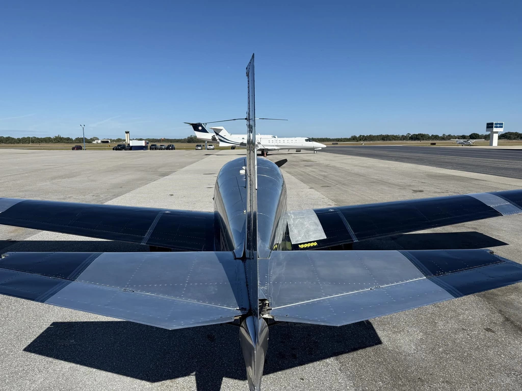 Rear view of a black and silver Mooney M20 aircraft, N5758Q, parked on an airfield with a white jet in the background.