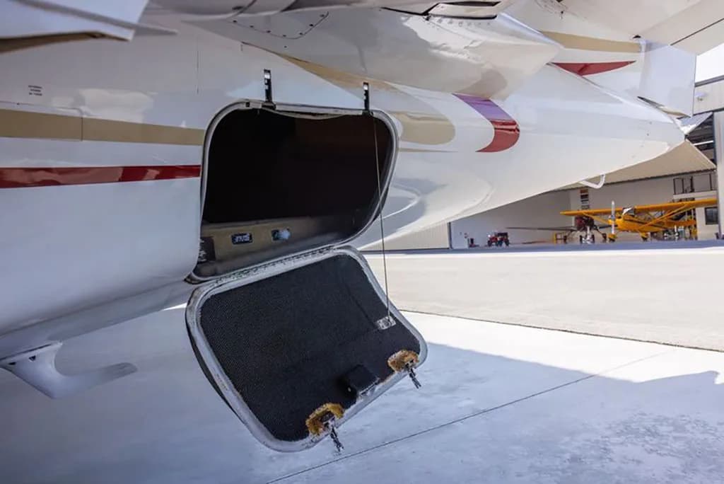 The rear fuselage of a white business jet with red and gold stripes, showing an open baggage compartment on an airport apron.