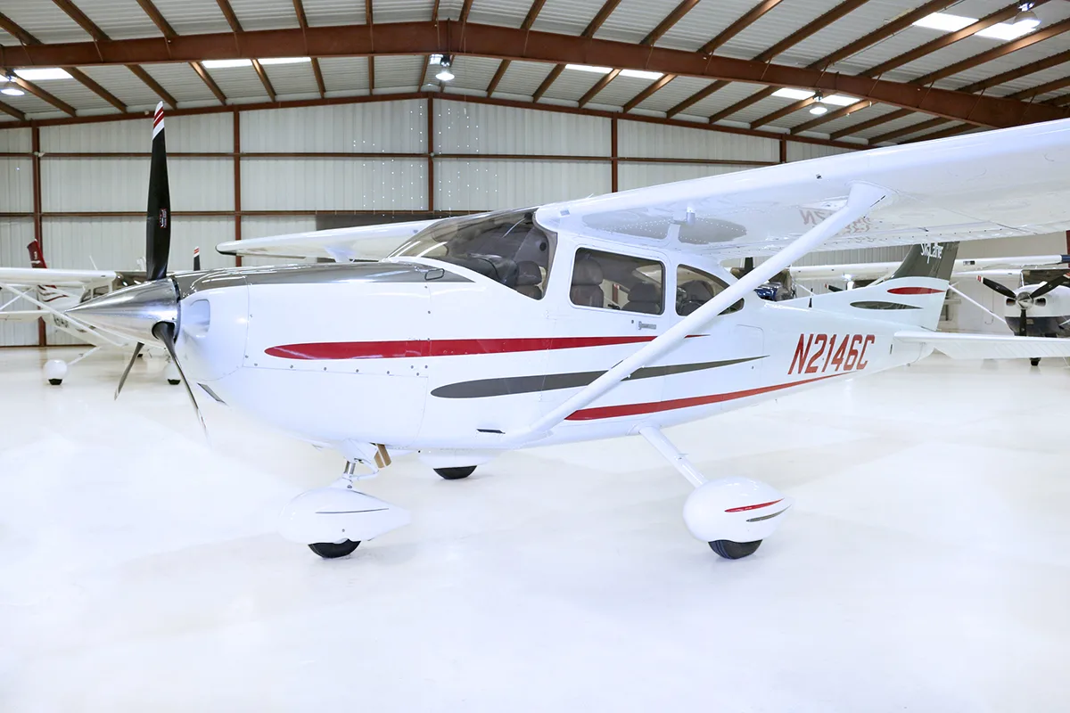 A white Cessna 182 Skylane with red and grey stripes, registration N2146C, parked in a hangar.