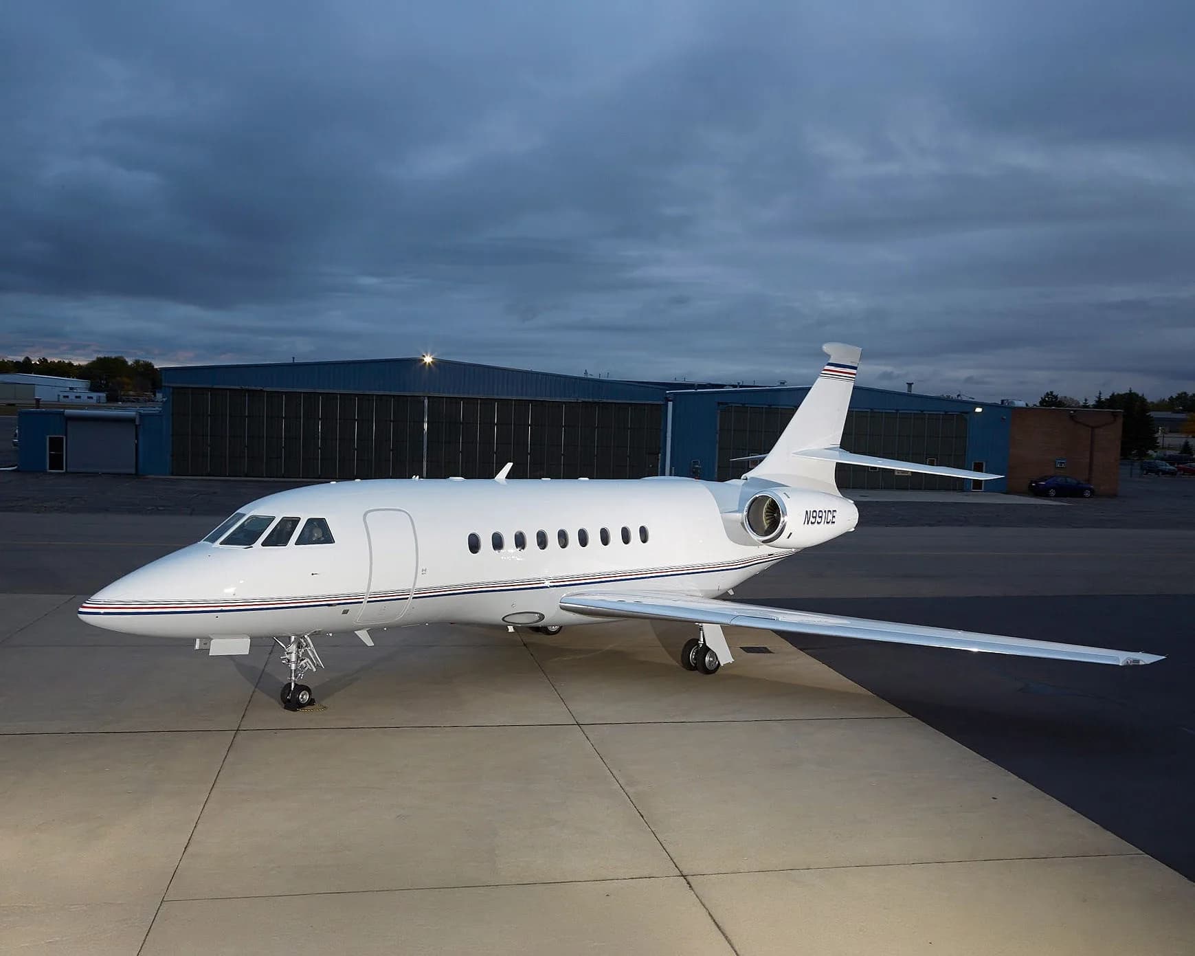 A white Dassault Falcon 900EX aircraft with blue and red stripes on the tail and fuselage, parked on an tarmac at dusk with hangars in the background.