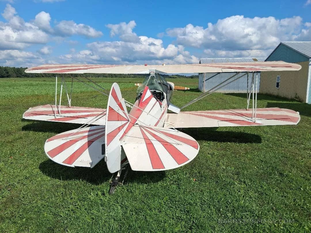 The rear view of a white biplane with red radial stripes on its tail and wings, parked on green grass under a cloudy sky.
