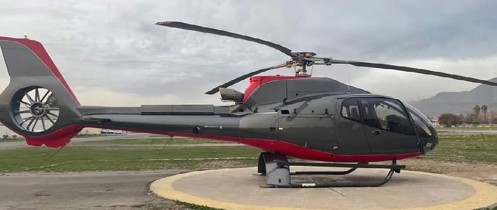 A dark grey and red helicopter is parked on a circular helipad with a grassy field and mountains in the background.