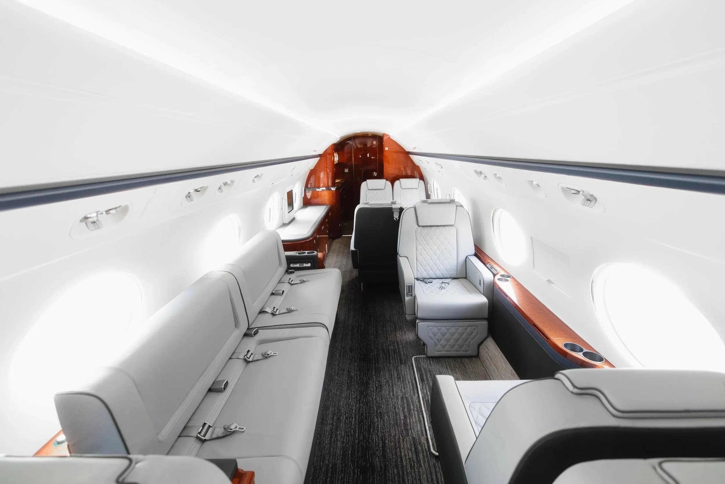 A wide view of the business jet's cabin, showing light grey seating, a divan, and dark wood cabinetry.