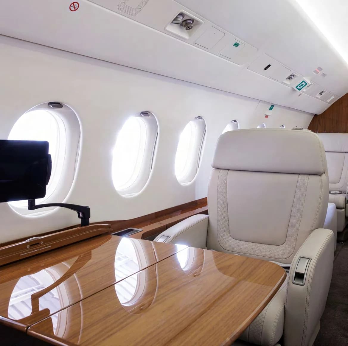 A side view of the business jet cabin featuring light grey leather seats, a polished wooden table, and multiple windows.