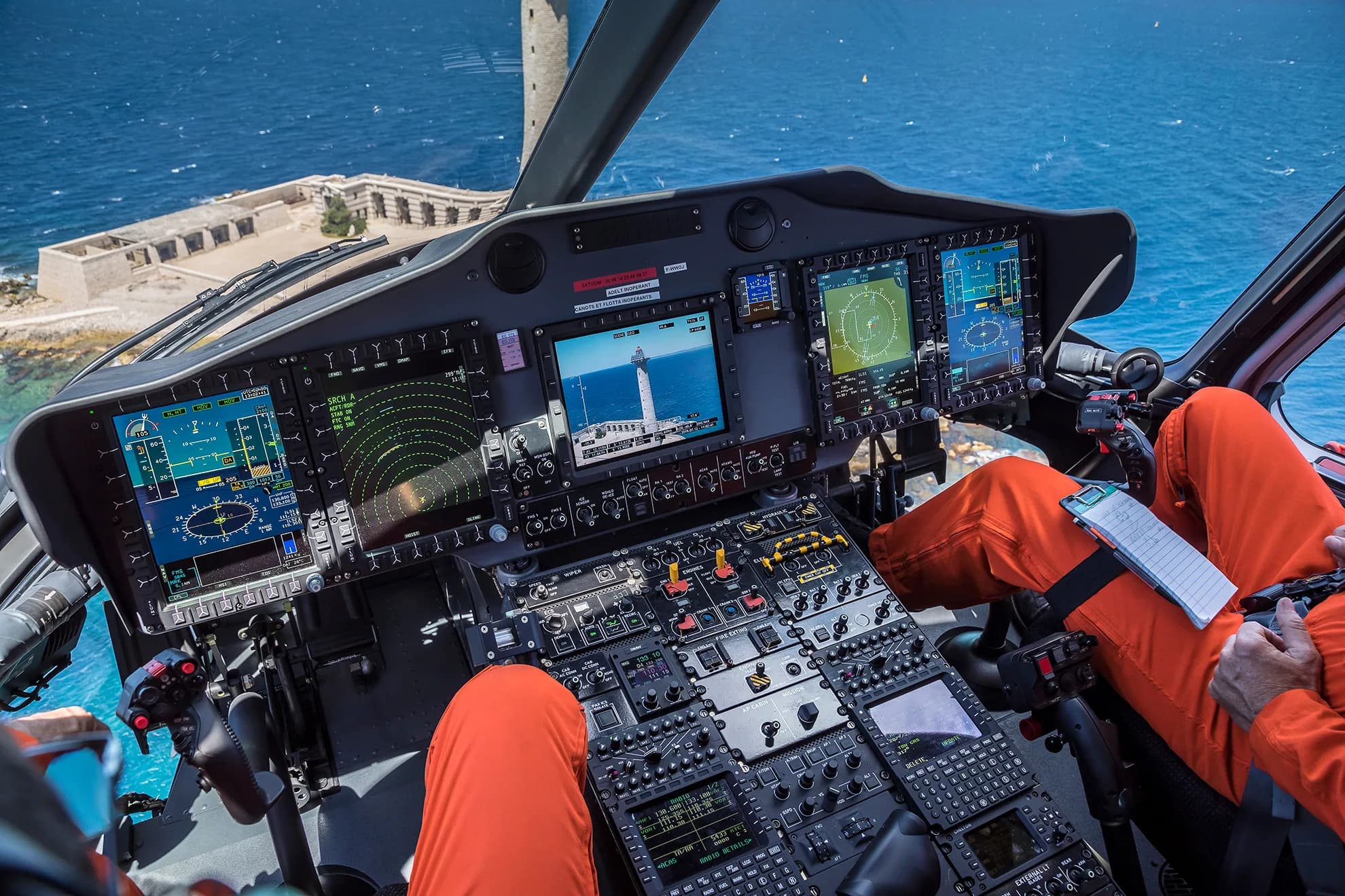 The cockpit of a helicopter with multiple display screens, showing a lighthouse on one, and a view of the sea and a coastal building outside.