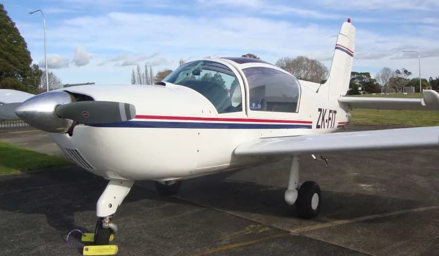 A white single-engine propeller aircraft with blue and red stripes, registration ZK-FIT, parked on an airfield tarmac.