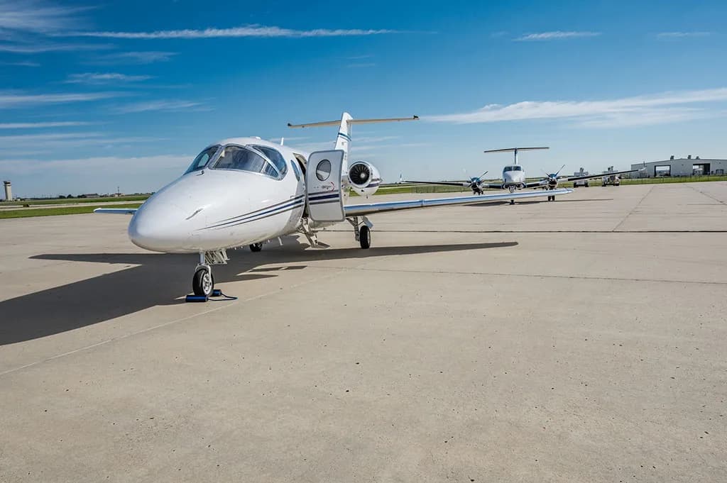 A white business jet with blue stripes and its entry door open, parked on an airport tarmac with other aircraft in the background.
