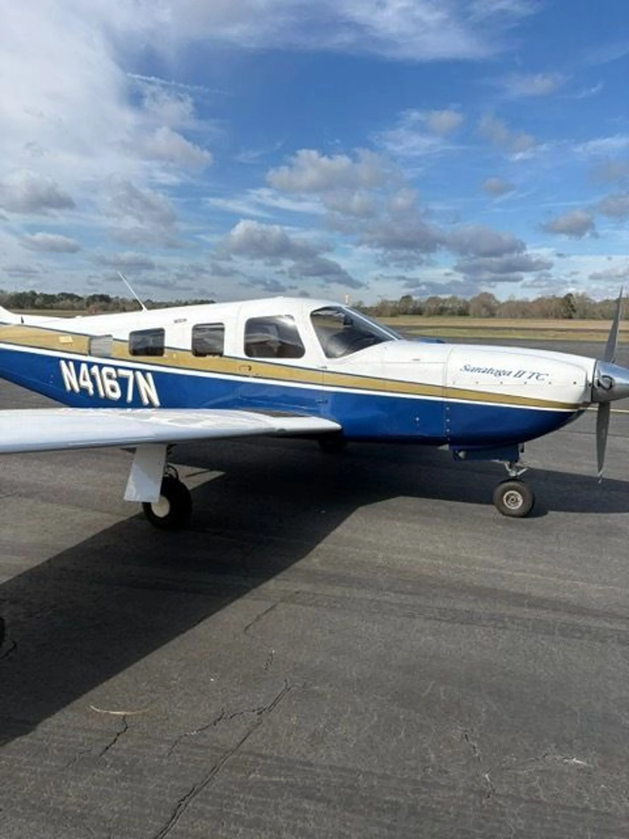 A side view of the Piper Saratoga II TC aircraft, N4167N, with white, blue, and gold paint, parked on an asphalt ramp.