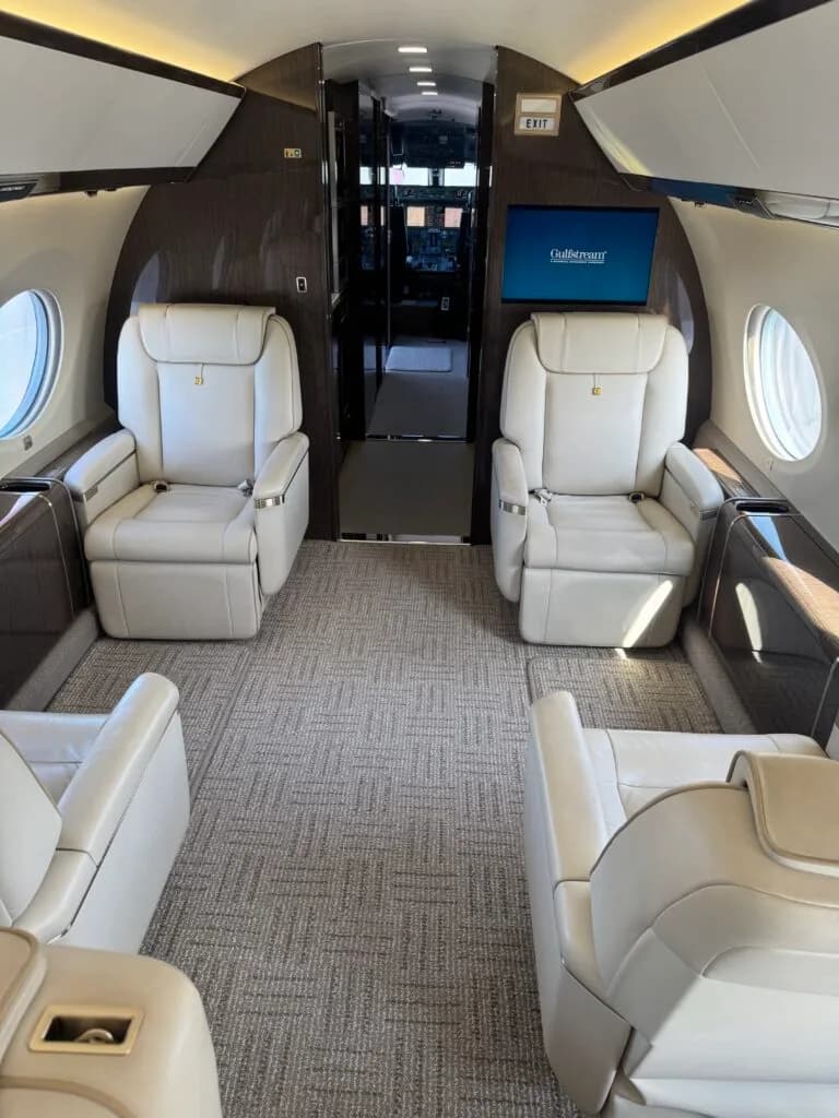 The luxurious interior of a Gulfstream cabin with light-colored leather seats, dark wood paneling, and a screen displaying 'Gulfstream' at the front.