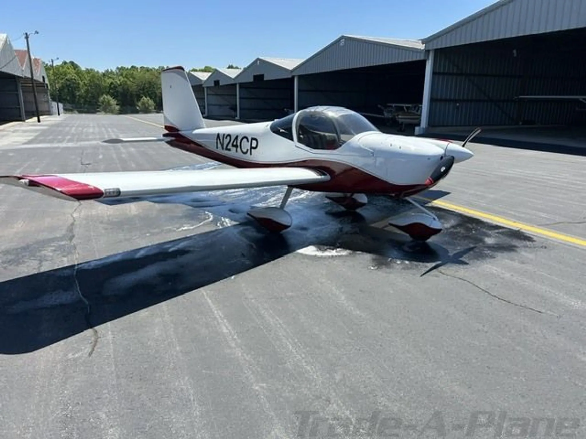 A white and red Van's RV-12 aircraft, N24CP, is parked on a tarmac with hangars in the background.