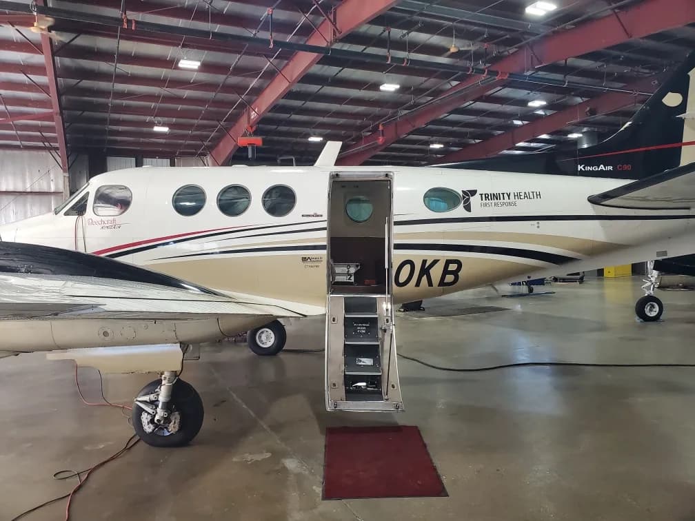 A white and tan Beechcraft King Air 90 turboprop aircraft with an open airstair door, parked in a hangar, viewed from the side.