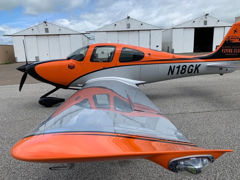 Three-quarter front view of a Cirrus SR20 aircraft, highlighting the orange, black, and silver paint scheme and wingtip light.