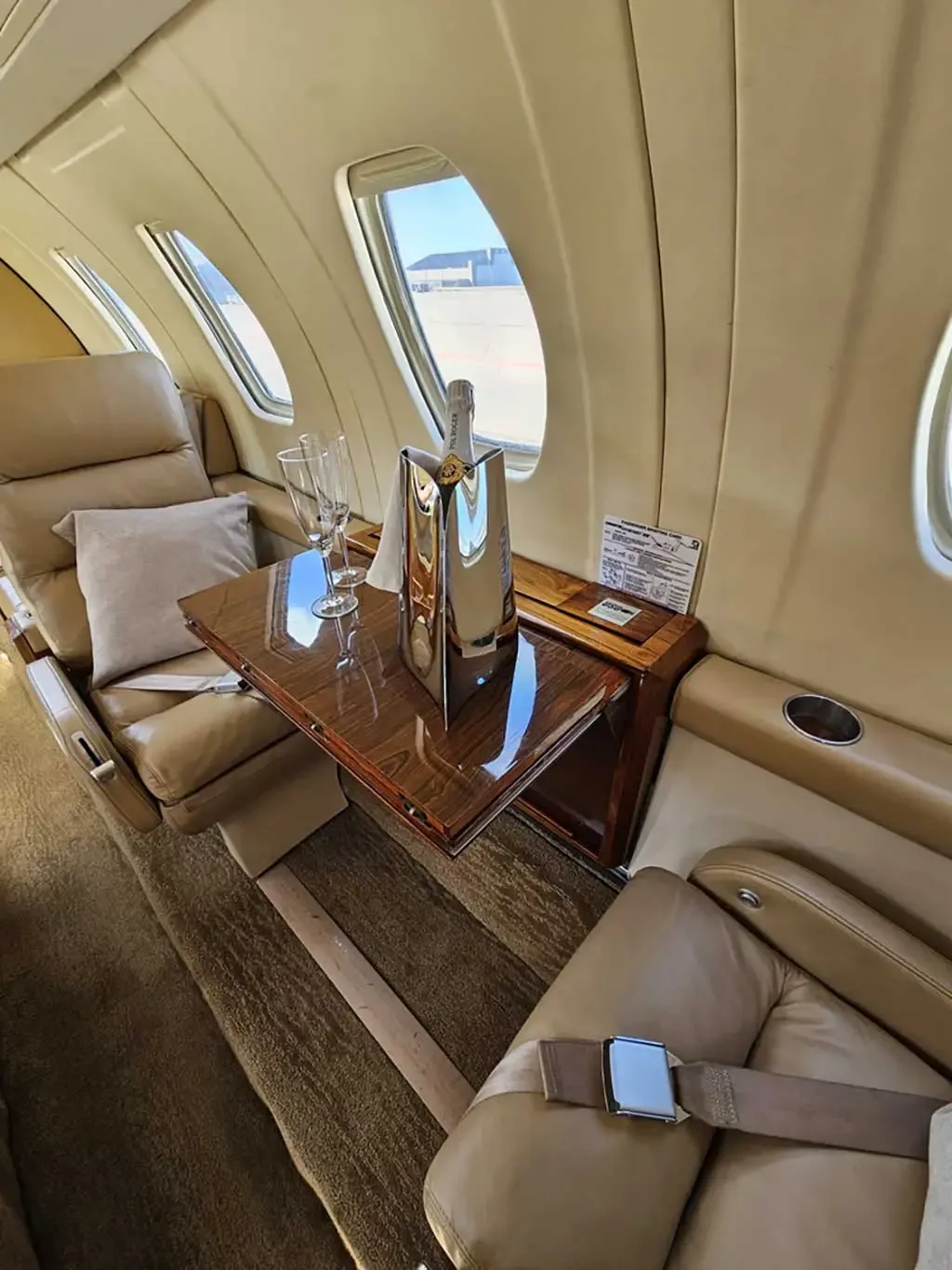 A close-up view of the private jet's interior, showing tan leather seats, a polished wooden table with champagne, and windows.