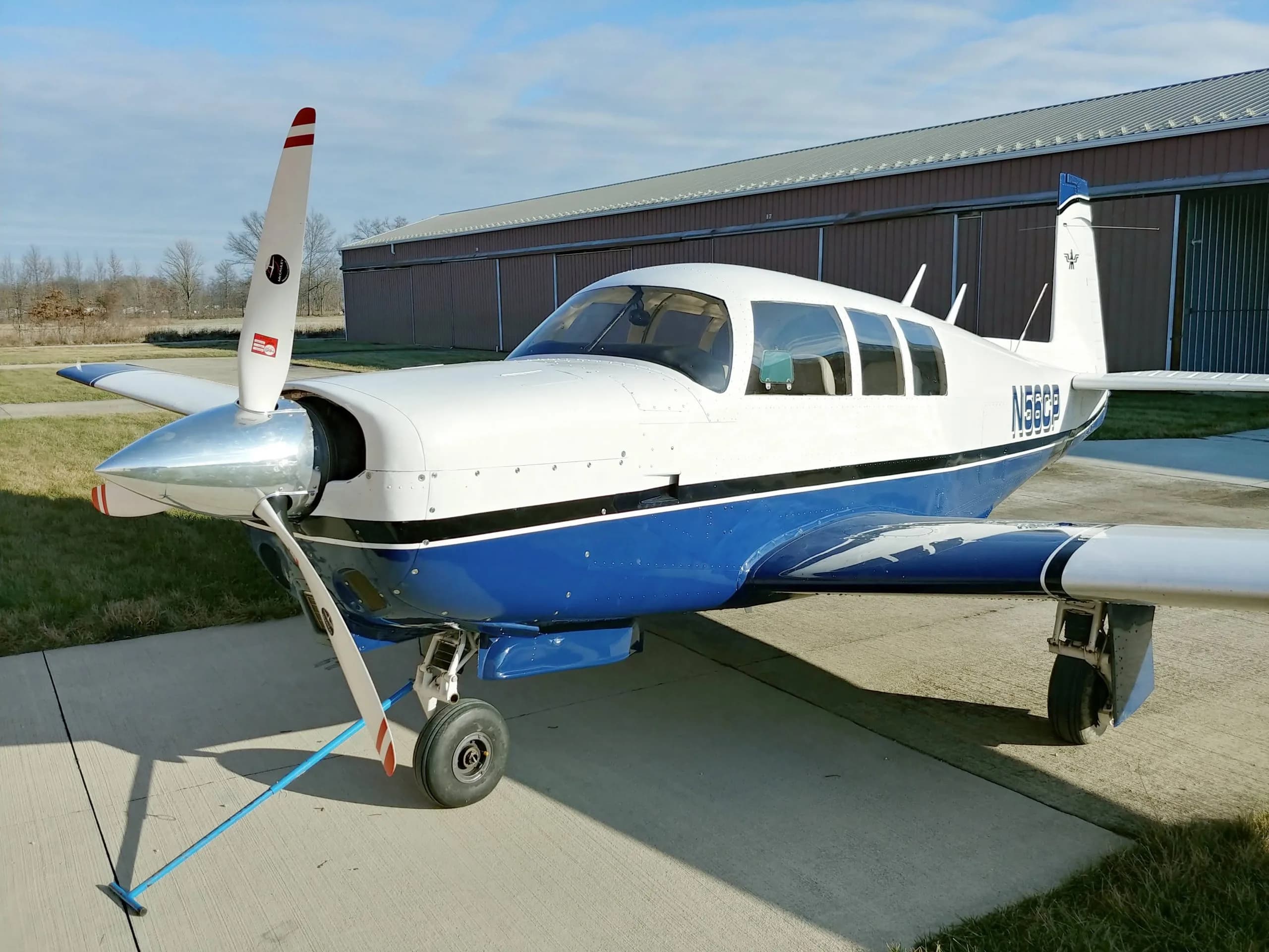 A blue and white Mooney aircraft, registration N56CP, is parked on the tarmac in front of a hangar.