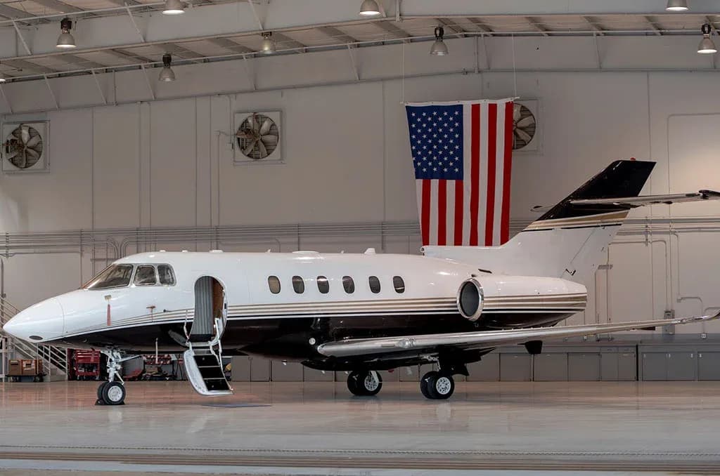 A white and black private jet with its main cabin door open and stairs extended, parked in a brightly lit hangar with an American flag.
