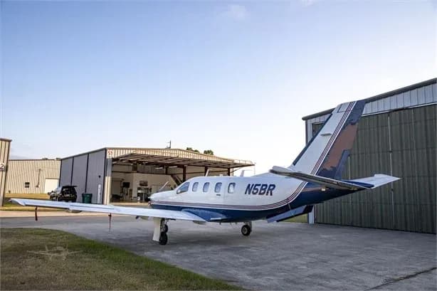 A white and blue single-engine turboprop aircraft, N5BR, is parked on an asphalt ramp with hangars in the background under a clear sky.
