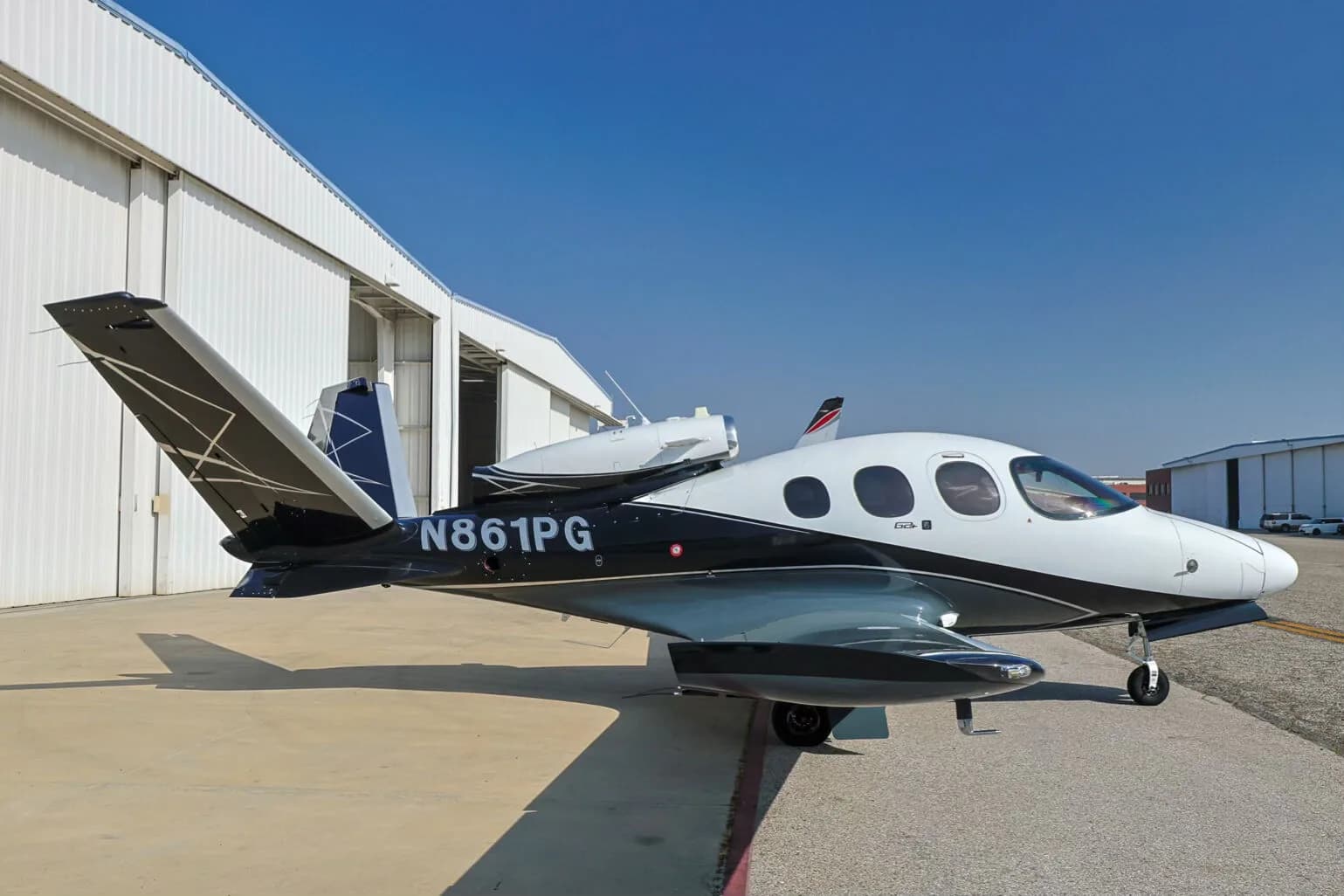 A Cirrus Vision Jet (N861PG) in white, black, and grey paint scheme parked on an tarmac next to a hangar.